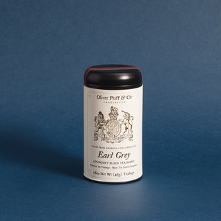 A cylindrical metal tea tin for Oliver Pluff & Co. Earl Grey, featuring a white label with black text and crest, a black lid with a red band, and a dark base, set against a textured blue background.