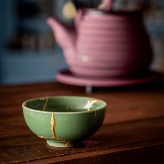 Green ceramic bowl with kintsugi repair on a wooden surface, blurred pink teapot in the background.