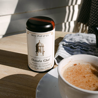 Oliver Platt & Co. Masala Chai tea canister with a steaming cup of tea on a wooden surface.
