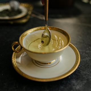 Gold-rimmed teacup with a spoon pouring liquid onto a dark surface