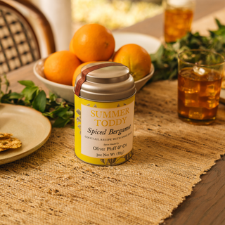 Tin of 'Summer Toddy' spiced bergamot cocktail mix on a table with oranges and glasses.