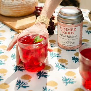 Person holding a glass of red drink next to a can labeled 'Summer Toddy Spiced Hibiscus' on a patterned tablecloth.